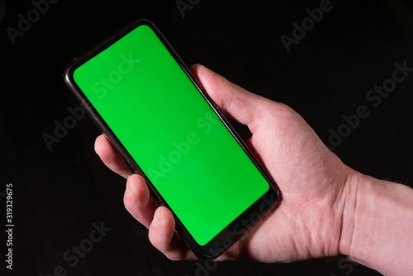 Fototapeta Male hand holds a black phone with a green screen on a dark background.