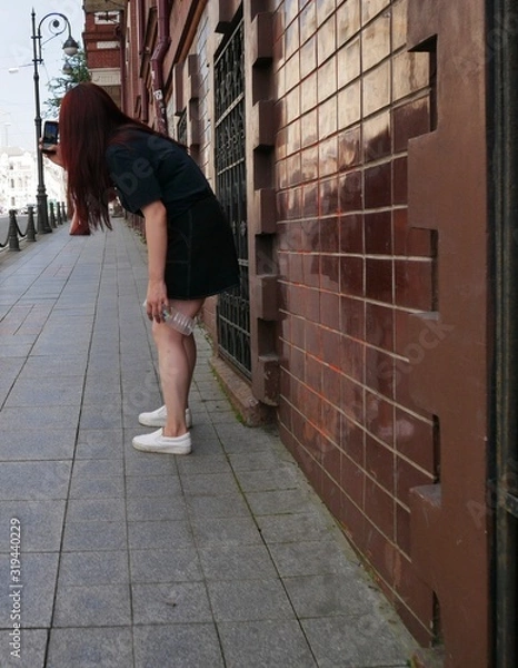 Fototapeta A girl with dark brown hair in a black t-shirt and a black mini skirt with a bottle of water in her hands takes photos of a city street with a smartphone on a Sunny summer day.