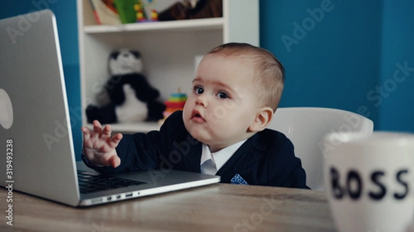 Obraz Photo of adorable serious baby boss in business suit typing on keyboard of the laptop while sitting in the modern office. Success, happy childhood, cuteness. Child’s portrait. Close up view