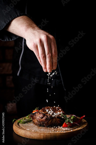 Obraz The concept of cooking meat. The chef cook salt on the cooked steak on a black background, a place under the logo for the restaurant menu. food background image, copy space text