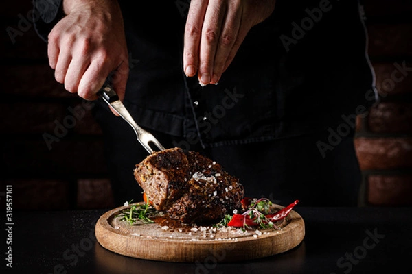 Fototapeta The concept of cooking meat. The chef cook salt on the cooked steak on a black background, a place under the logo for the restaurant menu. food background image, copy space text