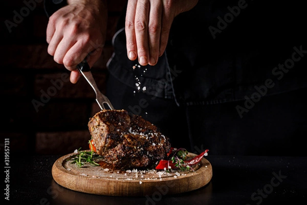 Obraz The concept of cooking meat. The chef cook salt on the cooked steak on a black background, a place under the logo for the restaurant menu. food background image, copy space text