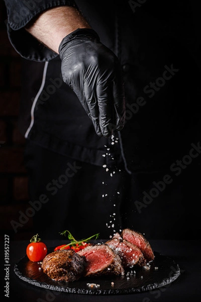 Obraz The concept of cooking meat. The chef cook salt on the cooked steak on a black background, a place under the logo for the restaurant menu. food background image, copy space text