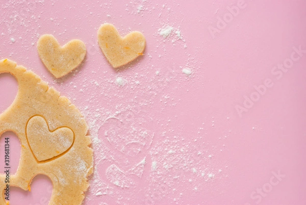 Fototapeta Cutting out heart-shaped cookies from flour-flavored dough on a light pink surface. Top view, the process of preparing homemade treats for Valentine's Day.