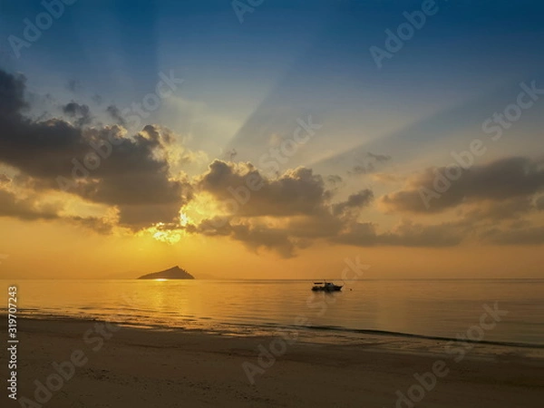 Fototapeta seaside of small boat floating in the sea with small island and sun rays with yellow sun light and cloudy sky background, sunrise at Ko Bulon Le, Stun Province, southern of Thailand.