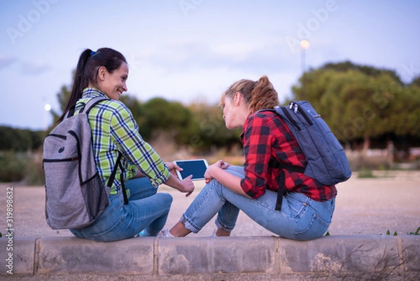 Fototapeta two young women students with backpack holding and looking at the tablet and smiling. Girl shows something on the screen.