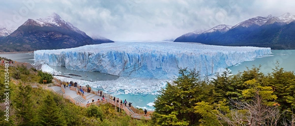 Fototapeta Panoramic view of Perito Moreno Glacier surrounded by green vegetation and snowy mountains, in El Calafate, Argentina, against a grey and cloudy sky.