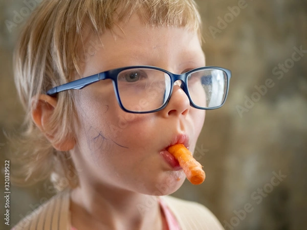 Fototapeta funny little blond boy in pink sweatshirt, big glasses and corset to correct posture eating fresh carrot. concept of vegetarianism and proper balanced nutrition for children