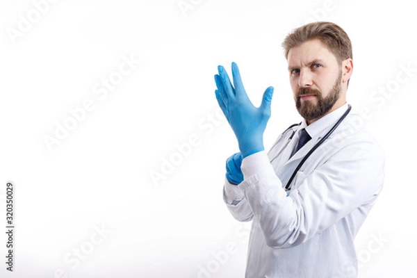 Obraz Confident bearded physician in white smock putting on blue medical gloves isolated white background copyspace