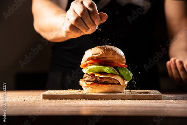Obraz Make it professional, home made hamburger made by white sesame bun, tomato slice, salad, cheese, grilled meat and onion on wooden tray wooden table with cooking decorate chef hand isolate in dark