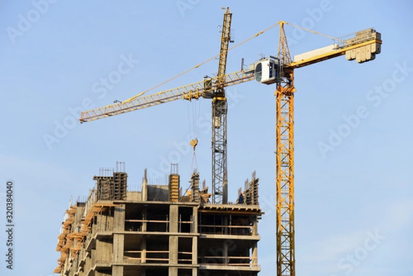 Fototapeta Tower cranes working at construction site against blue sky. Crane lifting a concrete bucket. Construction process of the new residential buildings. Transportation blocks and pouring of the cement mix