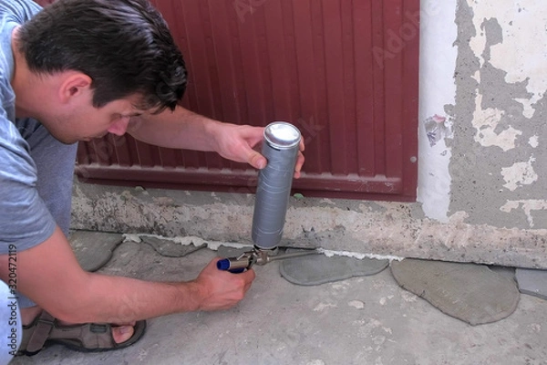 Fototapeta Young man worker fills crack between wall and cement floor with mounting foam in room under the battery. Repair and construction work of the house. Rough finish of the room.