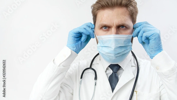 Fototapeta Potrait of practitioner putting on protective mask, having stethoscope, and blue surgical gloves while looking at camera isolated light background