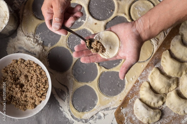 Fototapeta A man is making varenyky or dumplings, with meat, onions and bacon. Man is stuffing the dough with meat. Man in a black apron without face. Making dough by male hands at bakery Stone table close-up ph