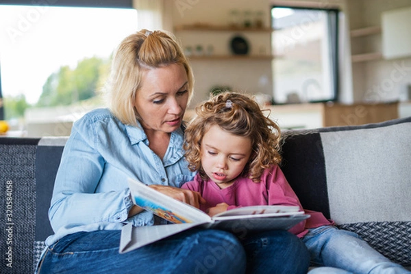 Fototapeta A cute small girl with mother on sofa indoors at home, reading book.