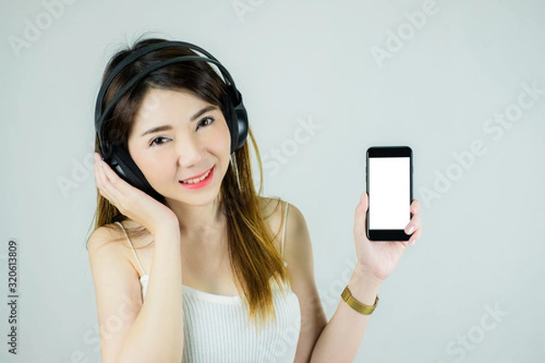 Obraz Portrait of beautiful asian woman with headphones and mobile device. Showing empty white screen. Caucasian female model isolated on white background.