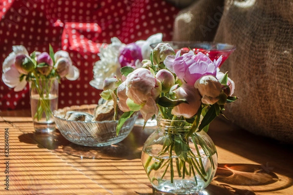 Fototapeta Beautiful little bouquet of undiscovered white pink peonies against the background of a red drink in a can. Buds of flowers stand in a glass little jar on the table in the sunlight, casting a shadow.