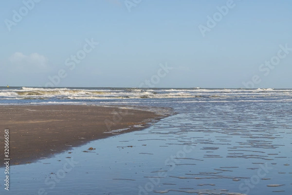 Fototapeta Dutch coastline. The sea surf at the sand and waves coming in with sea foam. Blue sky on a winter day.