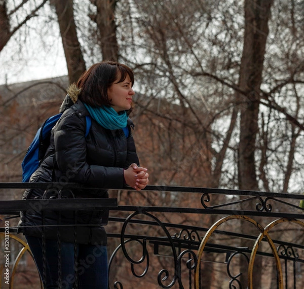 Fototapeta A young girl stands on a bridge in a city park on a weekend, sunny spring day. She enjoys the awakening of nature after winter and the warm sun. Early spring in the city.