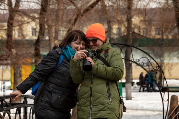Fototapeta Two girls take pictures and walk in a city park on a sunny day in early spring. A girl photographer takes a picture of a model and then watches the photos together on the camera.