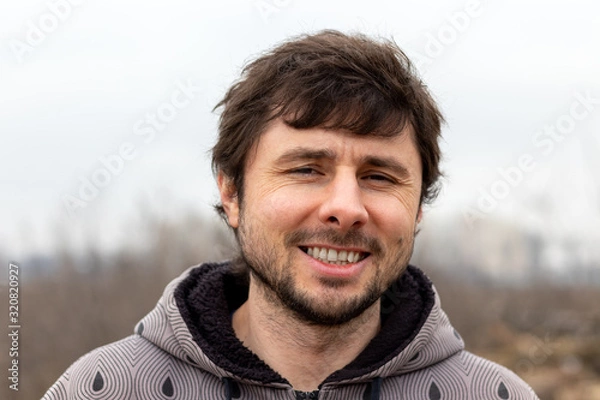Fototapeta A handsome happy smiling bearded man outside against a blurred background. Man in a jacket with a hood