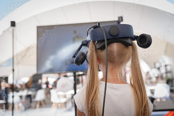 Fototapeta A little girl in a virtual reality helmet stands alone in front of a TV in an entertainment center