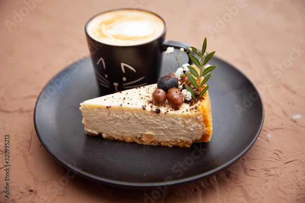 Fototapeta A black cup with a coffee is on a black plate near a piece of cake on which there are nuts and fruits