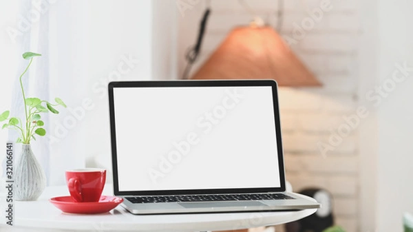 Fototapeta Computer laptop with white blank screen putting together with potted plant and coffee cup on the modern table with comfortable living room as background.
