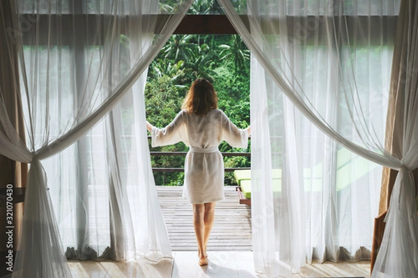 Fototapeta A woman in a bathrobe opens the curtains in deluxe Bali hotel room overlooking the terrace and tropical trees.Woman is awoke and standing before window. Girl is opening curtains and meeting sunrise