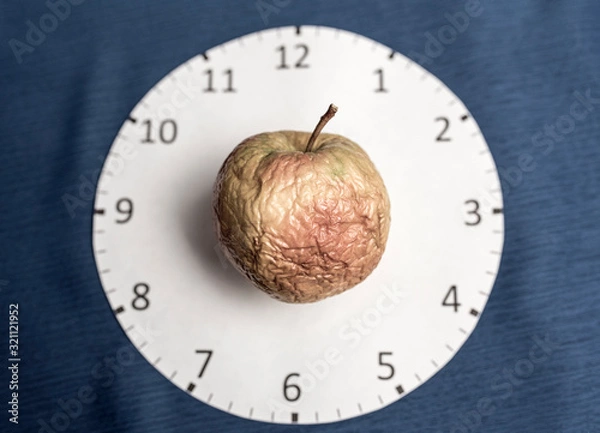 Fototapeta Clock face on a blue background and a wrinkled apple. Skin aging concept