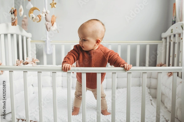 Fototapeta Beautiful little girl in a crib in a cozy light room in a sweater of fashionable shades. learns to stand at the support. cheerful little girl standing in the crib at home