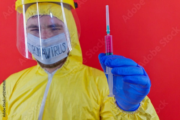 Obraz Man in yellow chemical protection suit and face protective mask coronavirus. Without glasses. Protective helmet. Hands in blue gloves. Syringe with red liquid.