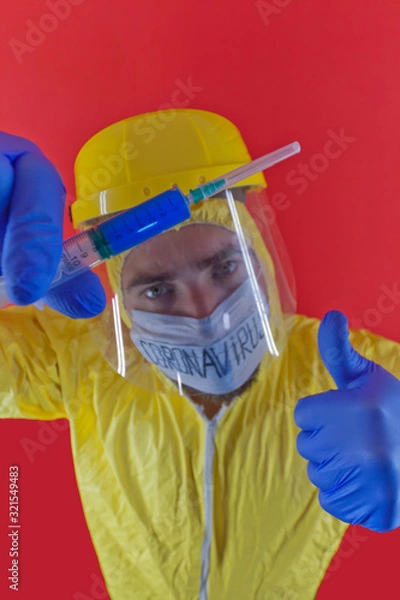 Obraz Man in yellow chemical protection suit and face protective mask  with the inscription `coronavirus`. Shoulder portrait. Red background. Without glasses. Protective helmet. Hands in blue gloves. 