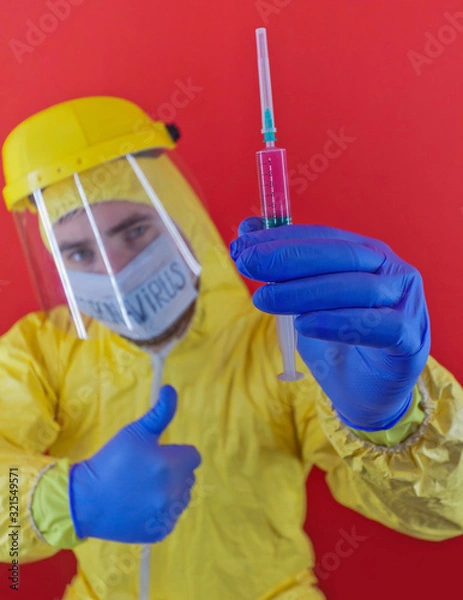 Obraz Man in yellow chemical protection suit and face protective mask  with the inscription `coronavirus`. Shoulder portrait. Red background. Without glasses. Protective helmet. Hands in blue gloves. 