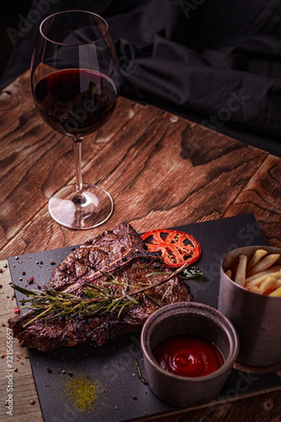 Fototapeta Cooked meat t-bone steak on serving board with garlic cloves, tomatoes, rosemary, spices and glass of red wine over rustic wooden background