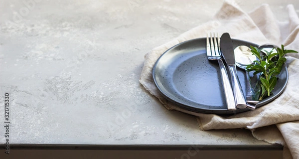 Fototapeta Elegant table setting with black ceramic plate, cutlery and canvas napkin on grey stone background. Space for text.