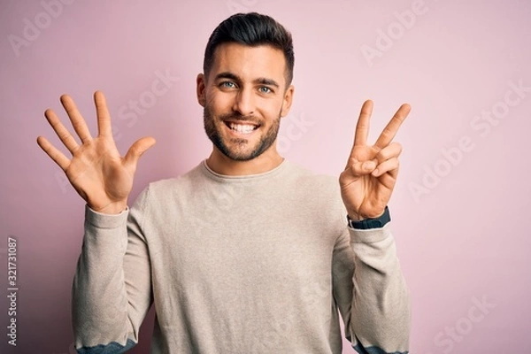 Fototapeta Young handsome man wearing casual sweater standing over isolated pink background showing and pointing up with fingers number seven while smiling confident and happy.