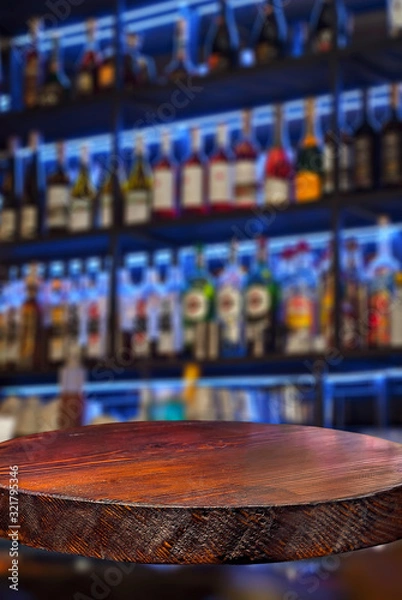 Fototapeta Wooden board on a background of bottles with alcohol. Old bar counter as layout for design. Workpiece for design. Empty place to advertise products. Blurred interior of the bar in the background.