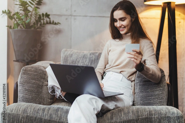 Fototapeta Girl with laptop sitting at home and making orders in online shops. Young woman doing payments with bank card for internet purchases. Debit and credit cards with cashback.