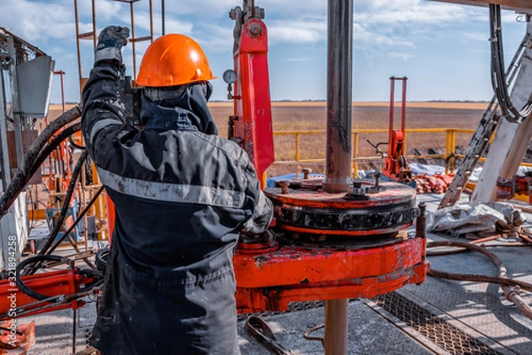 Fototapeta Work driller in red uniform, in helmet and goggles. He uses a hydraulic wrench to screw drill pipes to lower them into an oil well and continue drilling it. The concept of a working person.