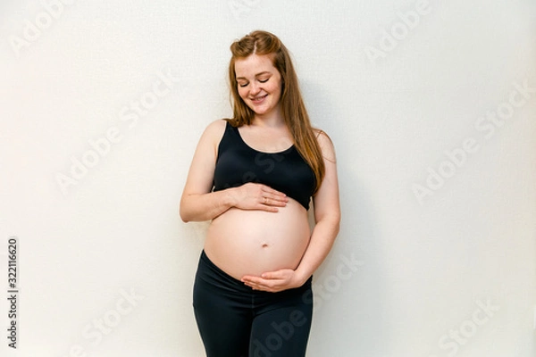 Fototapeta Image of pregnant woman in the black sport leggins and bra at thirty week (seven monthes) in the top and leggings, arms hugging her stomach. Pleasant expectation baby