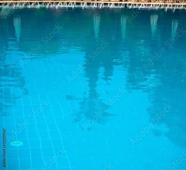 Fototapeta Outdoor pool. Through the water you can see the bottom of the pool. Palm trees and sun umbrellas are reflected in the water. Transparent water background with reflections.