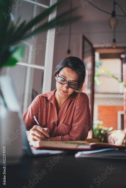 Obraz Young female administrative manager in eyeglasses making notes of information working at modern at office, People Business Lifestyle