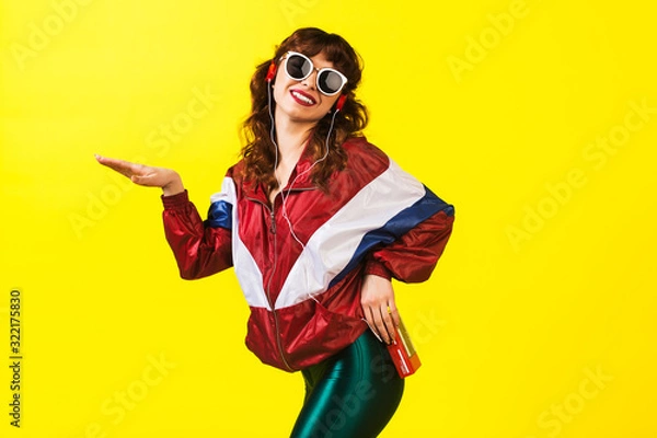 Obraz Cheerful gorgeous girl in clothes in the style of the 90s, with a vintage cassette player and headphones, in the studio on a white background. Music, emotions, fashion