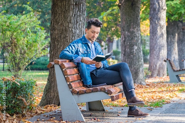 Fototapeta Handsome young man reading a book in a park. Portrait of a young man with a denim jacket and blue shirt reading a book outside. A guy reading a book in a park