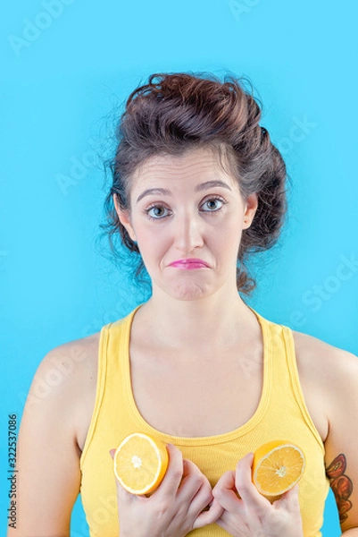 Fototapeta Beautiful brunette in yellow top holds lemons like tits over blue background, beauty concept