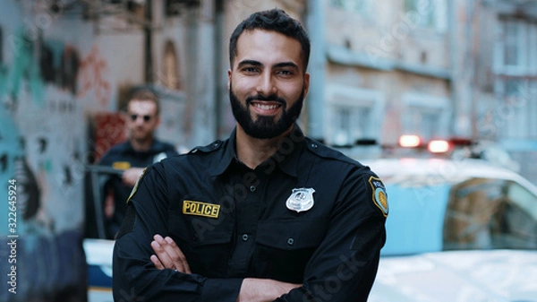 Obraz Uśmiechnięty młody człowiek policjanci stoją w pobliżu samochodu patrolowego spójrz na egzekwowanie kamer szczęśliwy policjant mundur policyjny bezpieczeństwo auto bezpieczeństwo komunikacja kontrola policjant portret zbliżenie zwolnione tempo