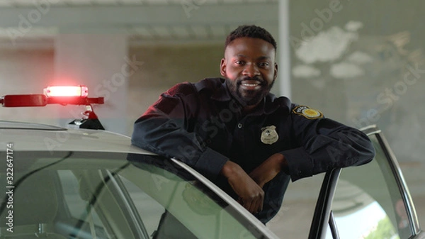 Obraz Smiling african american young man cops stand near patrol car look at camera enforcement happy officer police uniform auto safety security communication control policeman close up slow motion