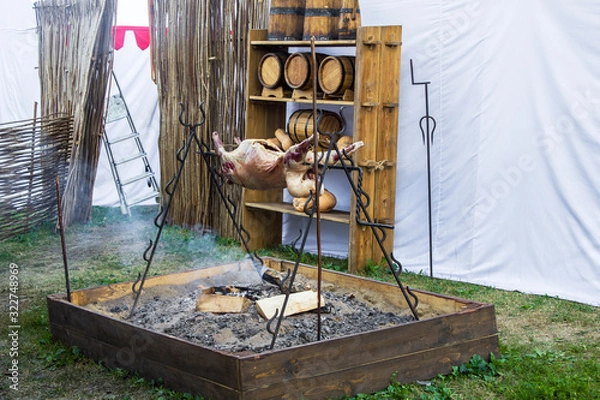 Fototapeta a large carcass of an animal is being cooked on a fire against the background of wooden old barrels and a tent