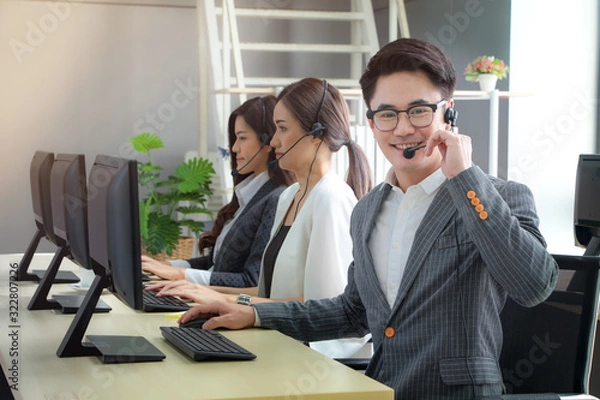Fototapeta Side view of handsome young asian operator man and woman in headsets using laptops while working in office. Male looking at camera. Concept of call center service for business.